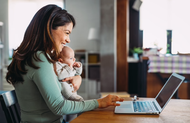 Person holding an infant while using a laptop at a table in a home office.