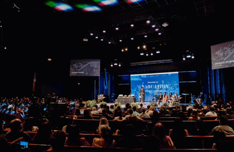 Wide view of a graduation ceremony in a large auditorium, with attendees seated facing a stage where speakers and graduates are gathered.