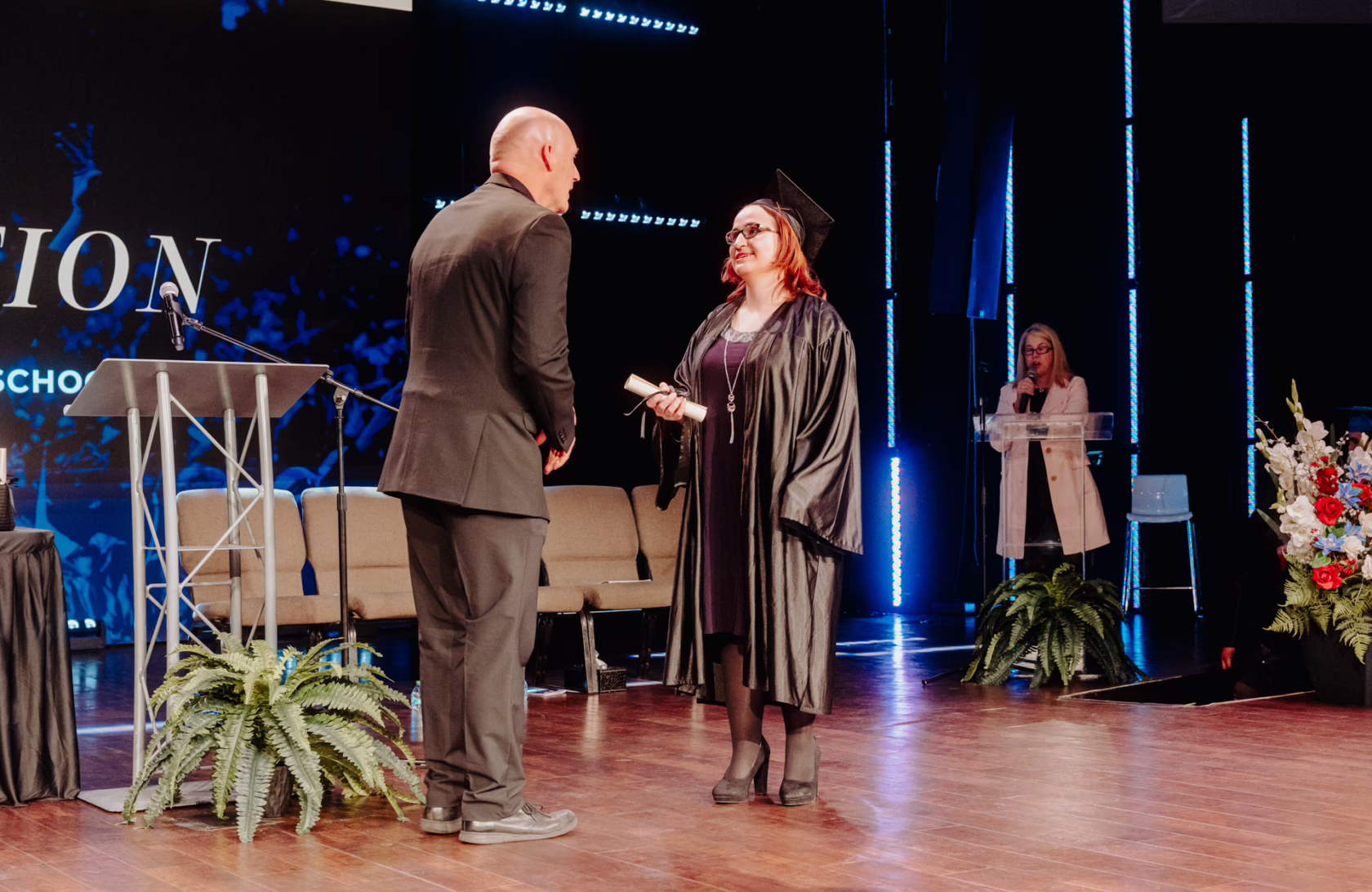 On a graduation stage, a graduate in a black cap and gown stands facing a person in a dark suit while holding a rolled diploma. A podium with a microphone and a potted plant sit nearby. In the background, a person speaks at a clear lectern, while blue vertical stage lights and a large screen with “Graduation 2026” text create a formal ceremony setting. Floral arrangements decorate the stage.