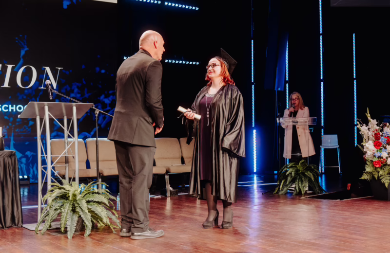 On a graduation stage, a graduate in a black cap and gown stands facing a person in a dark suit while holding a rolled diploma. A podium with a microphone and a potted plant sit nearby. In the background, a person speaks at a clear lectern, while blue vertical stage lights and a large screen with “Graduation 2026” text create a formal ceremony setting. Floral arrangements decorate the stage.