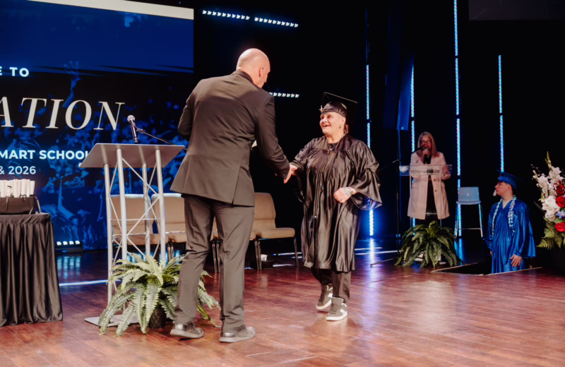 On a graduation stage, a person in a cap and gown walks forward to shake hands with a person in a dark suit near a podium with a microphone. Behind them, a large screen displays “Graduation 2026.” Stage lighting in blue tones lines the background, while a host stands at a clear lectern to the side and another graduate in a blue gown waits nearby. A table with stacked items and floral arrangements decorates the stage.
