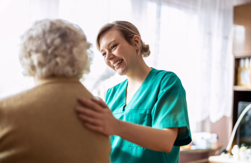 A healthcare worker in green scrubs stands indoors near a window, placing a hand on the shoulder of an older person with short, light-colored hair who is facing away from the camera, in a softly lit home-like setting with shelves and household items in the background.