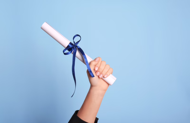 Hand holding a rolled-up white diploma tied with a blue ribbon against a light blue background.