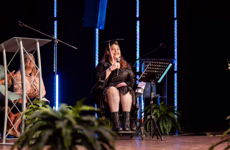 High school graduate in cap and gown speaking on stage at commencement ceremony using microphone, seated in wheelchair
