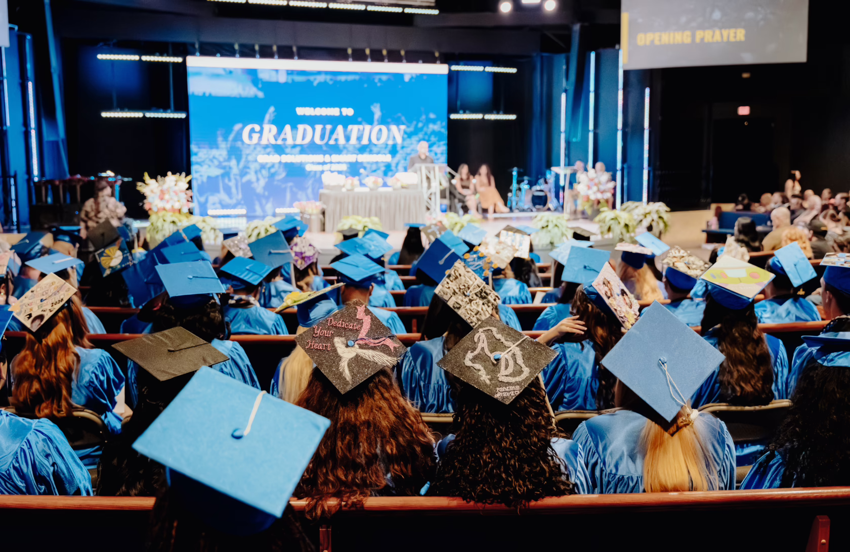 Rows of graduates in blue caps and gowns sit facing a stage during a high school graduation ceremony, with many decorated mortarboards visible in the audience.