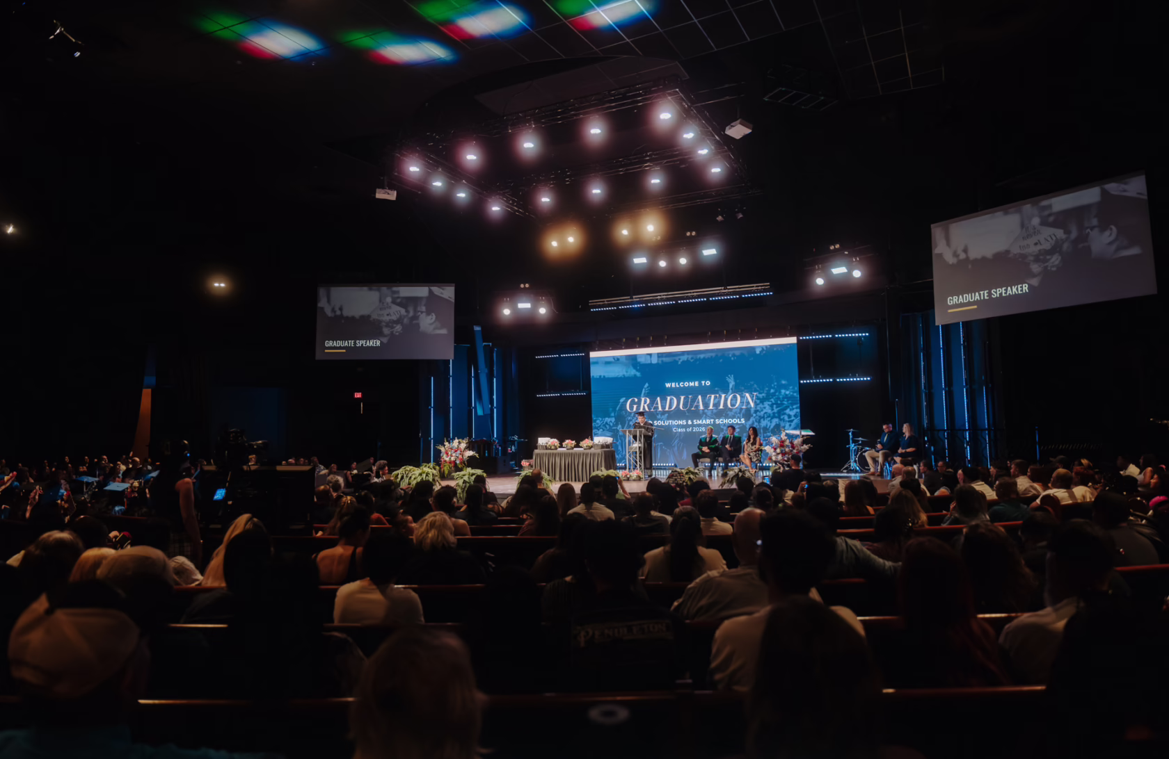 Wide view of a graduation ceremony in a large auditorium, with a speaker at a podium on a brightly lit stage, graduates and guests seated throughout the audience, and large screens displaying ceremony content.