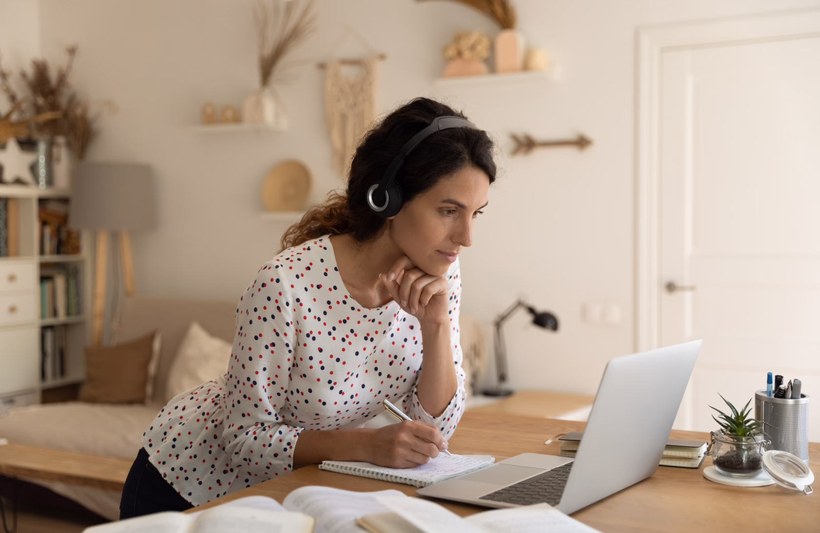 Adult learner studying online at home, taking notes while attending a virtual class on a laptop to earn a high school diploma.