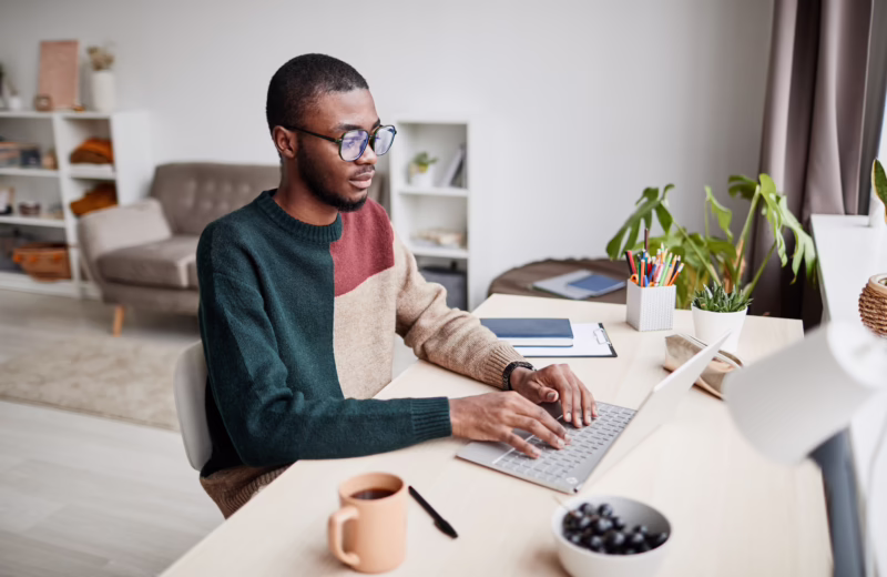 Person working on a laptop in a home study space, with notebooks, writing supplies, and a coffee mug on the desk while completing online learning activities.