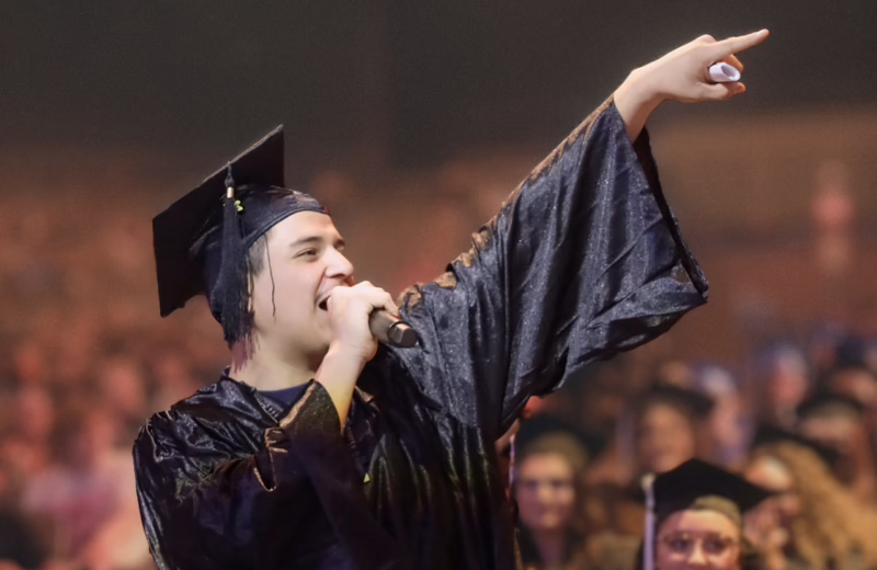 Graduate in a cap and gown speaks into a microphone during a graduation ceremony, gesturing toward the audience.