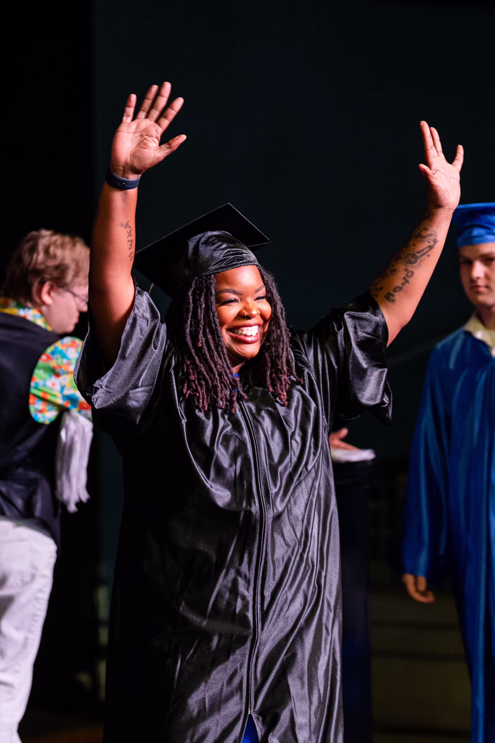 Graduate in cap and gown raising both hands while walking across a graduation ceremony stage.