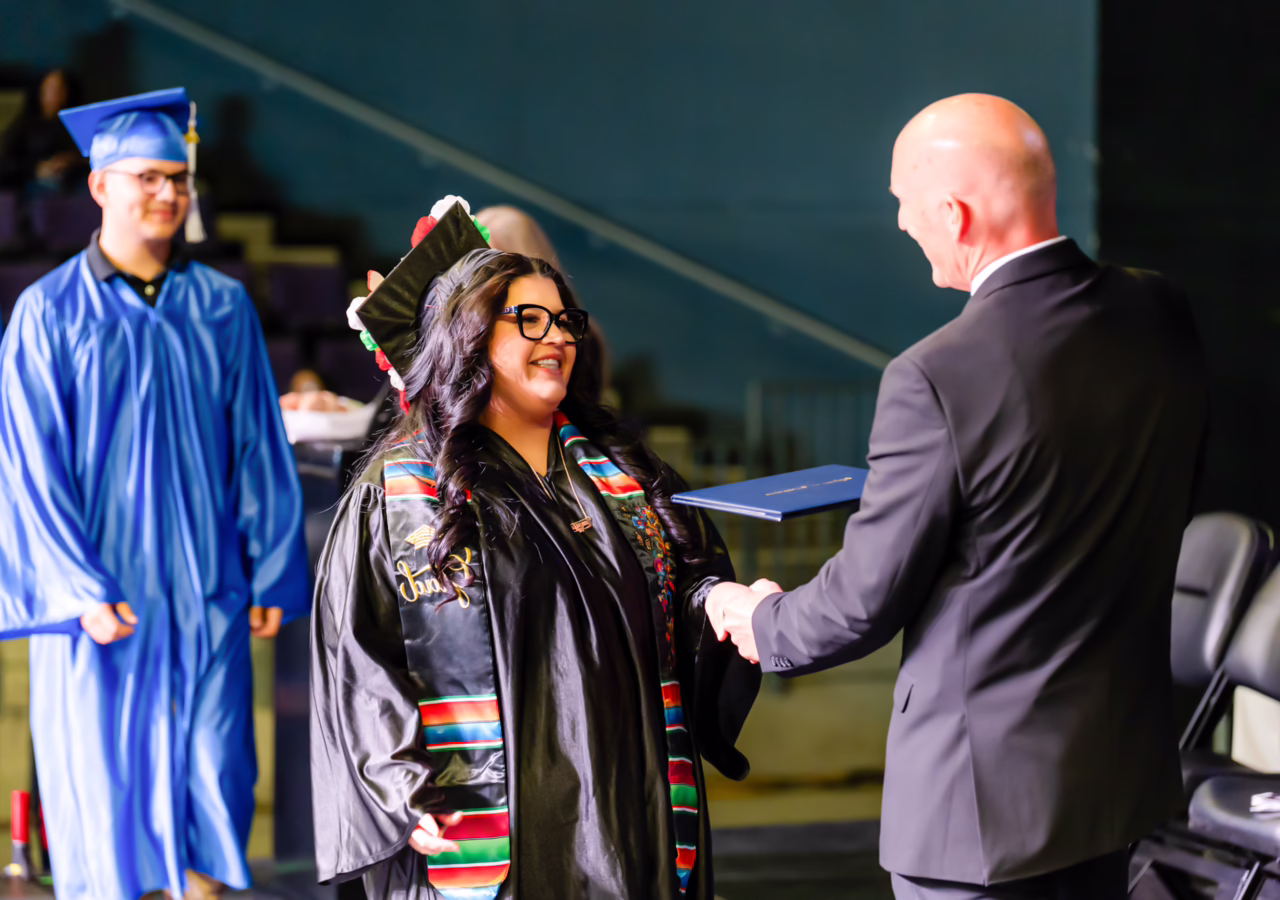 Graduate in a cap and gown shakes hands with a presenter while receiving a diploma during a graduation ceremony.