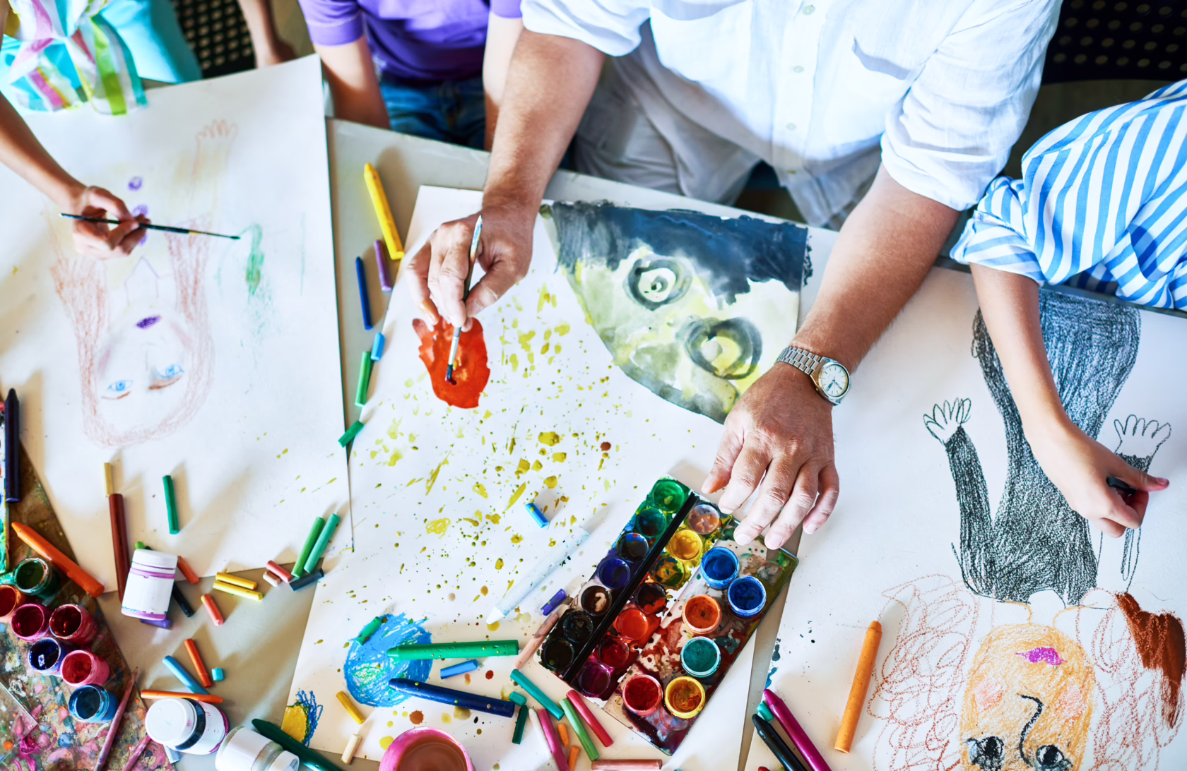 Overhead view of several people creating artwork on large sheets of paper using watercolor paints, brushes, and colorful drawing supplies.