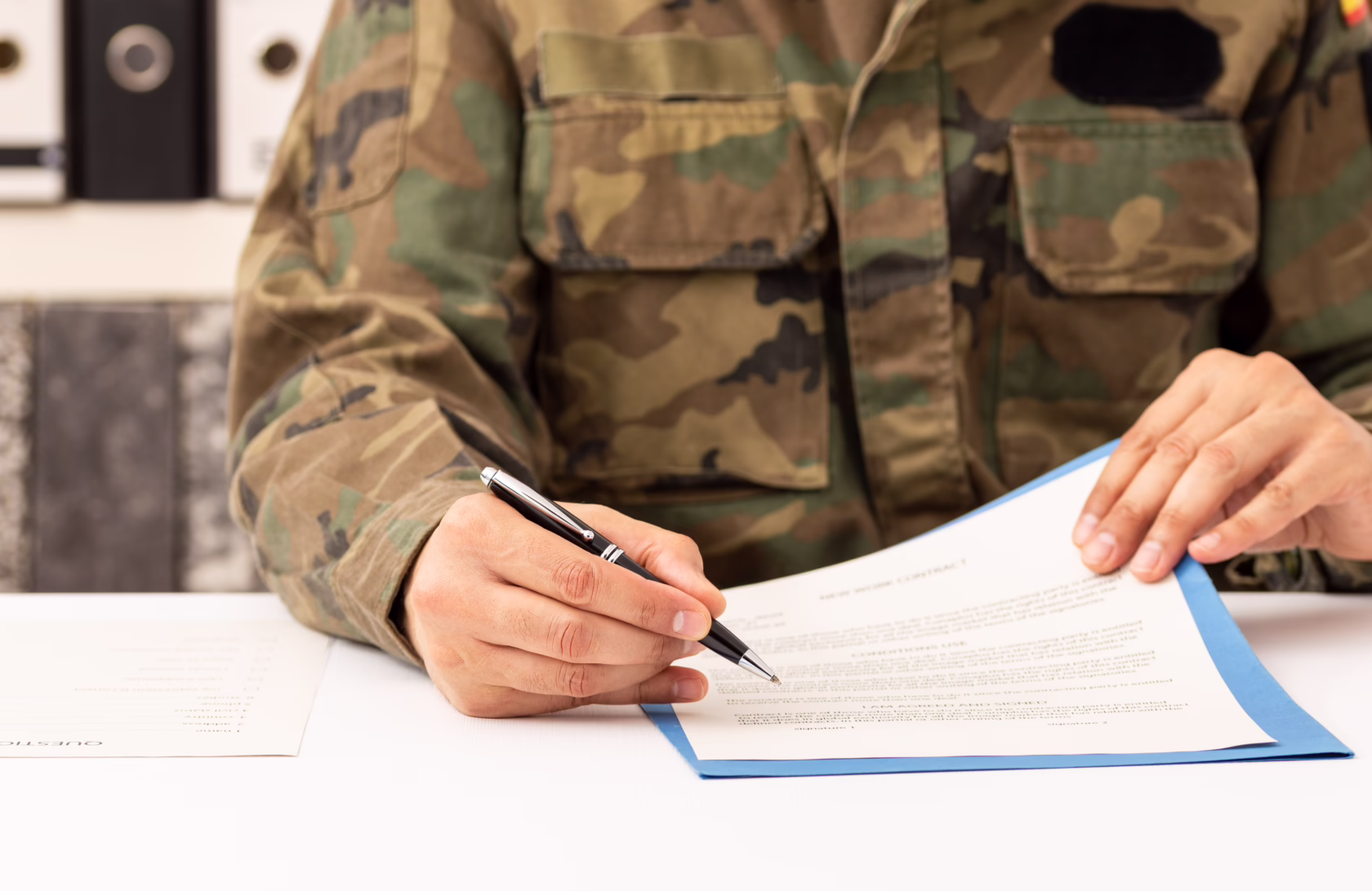 Military service member in camouflage uniform signing enlistment paperwork at a desk.
