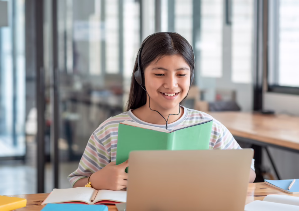 Person wearing headphones participates in online learning, reading a book while using a laptop at a desk.