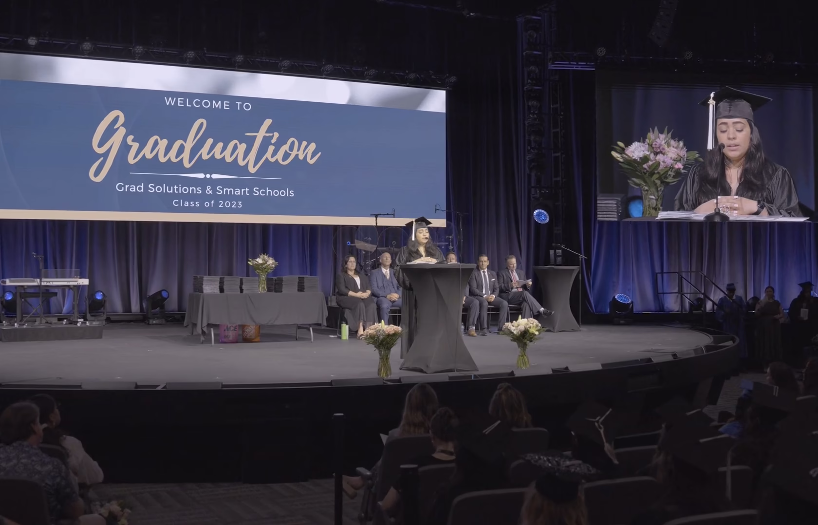 Graduate speaker addresses an audience from a podium during a graduation ceremony while a large screen displays commencement event information.