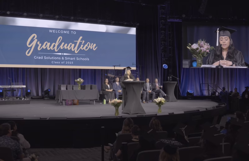 Graduate speaker addresses an audience from a podium during a graduation ceremony while a large screen displays commencement event information.