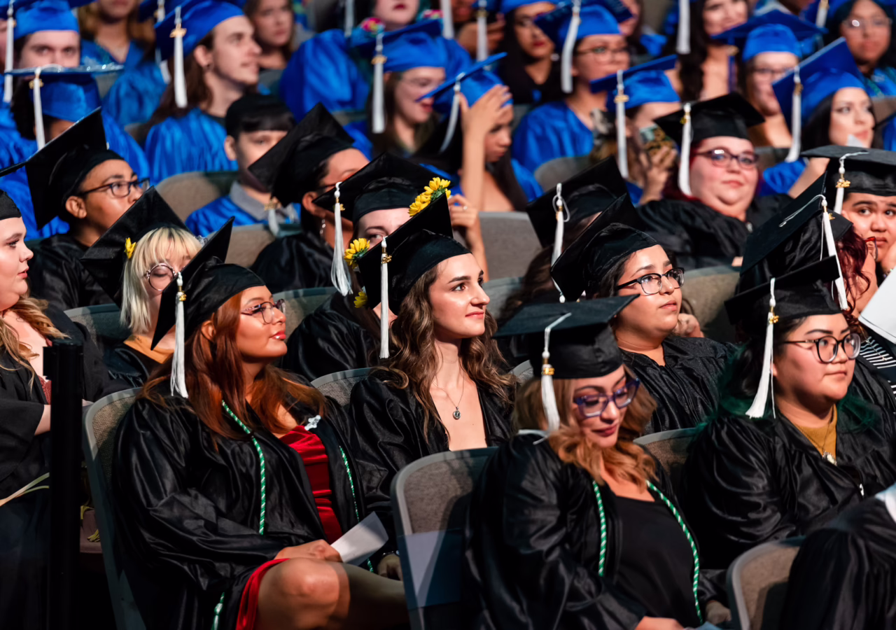 Graduates wearing caps and gowns sit together during a graduation ceremony, listening as the event takes place.