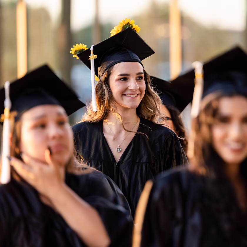 Graduates wearing black caps and gowns gather outdoors during a commencement ceremony, with one graduate in focus in the center of the group.