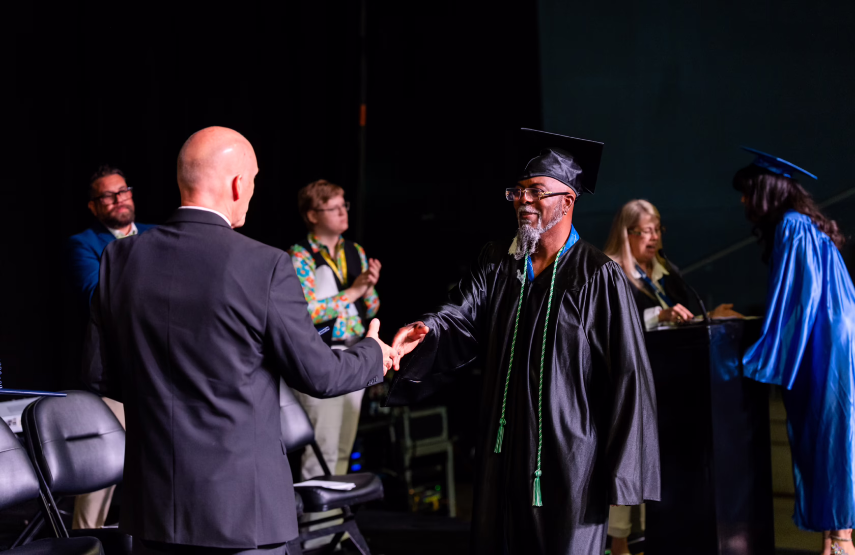 Adult graduate in cap and gown shakes hands on stage during a graduation ceremony after receiving a diploma.