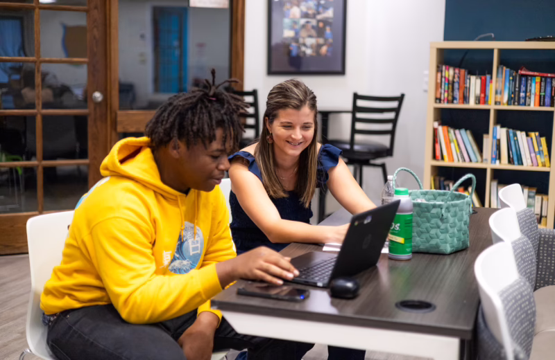 Two people sit at a table using a laptop in a learning or tutoring space, with bookshelves and study materials in the background.