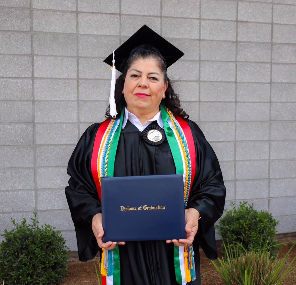 Graduate wearing a black cap and gown with colorful honor cords and stoles, holding a blue diploma cover with gold lettering while standing outdoors in front of a gray block wall.