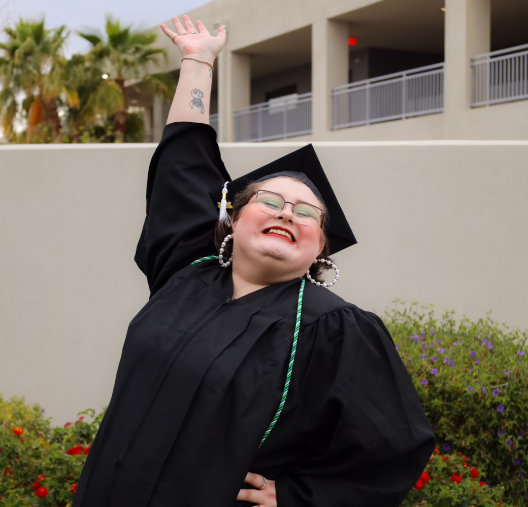 Graduate in a black cap and gown poses outdoors with one arm raised beside a landscaped campus building.