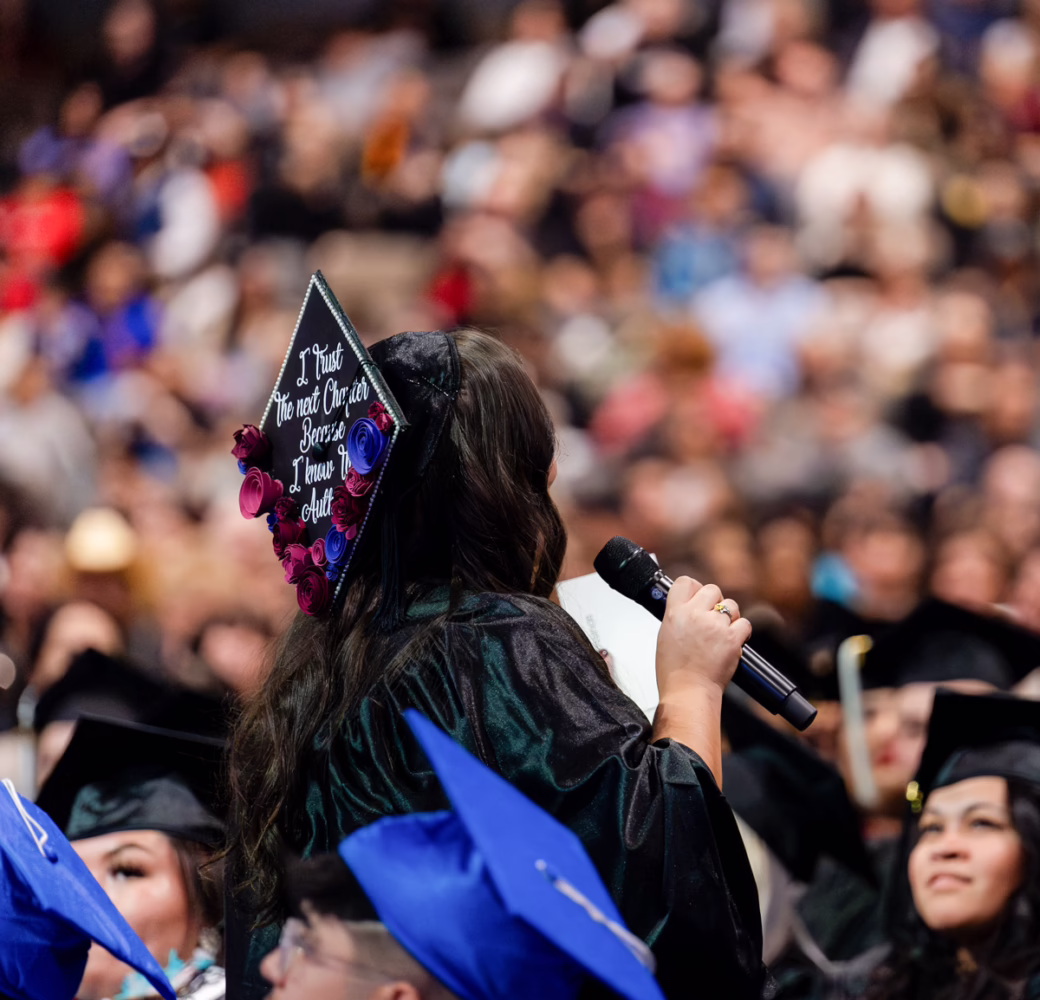 a graduate in a decorated cap and gown stands up and holds a microphone to thank friends and family during a commencement ceremony