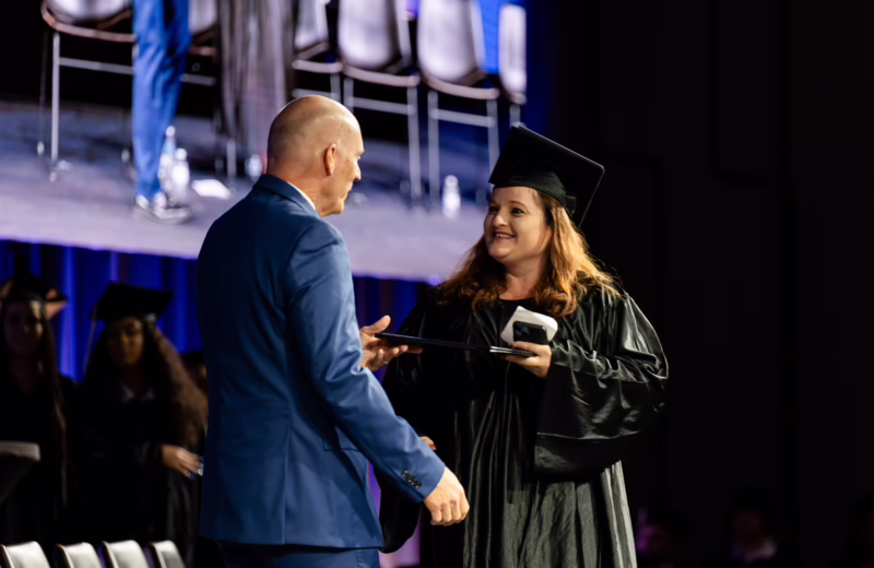 Graduate in a black cap and gown receives a diploma on stage during a graduation ceremony while shaking hands with a presenter in a blue suit.