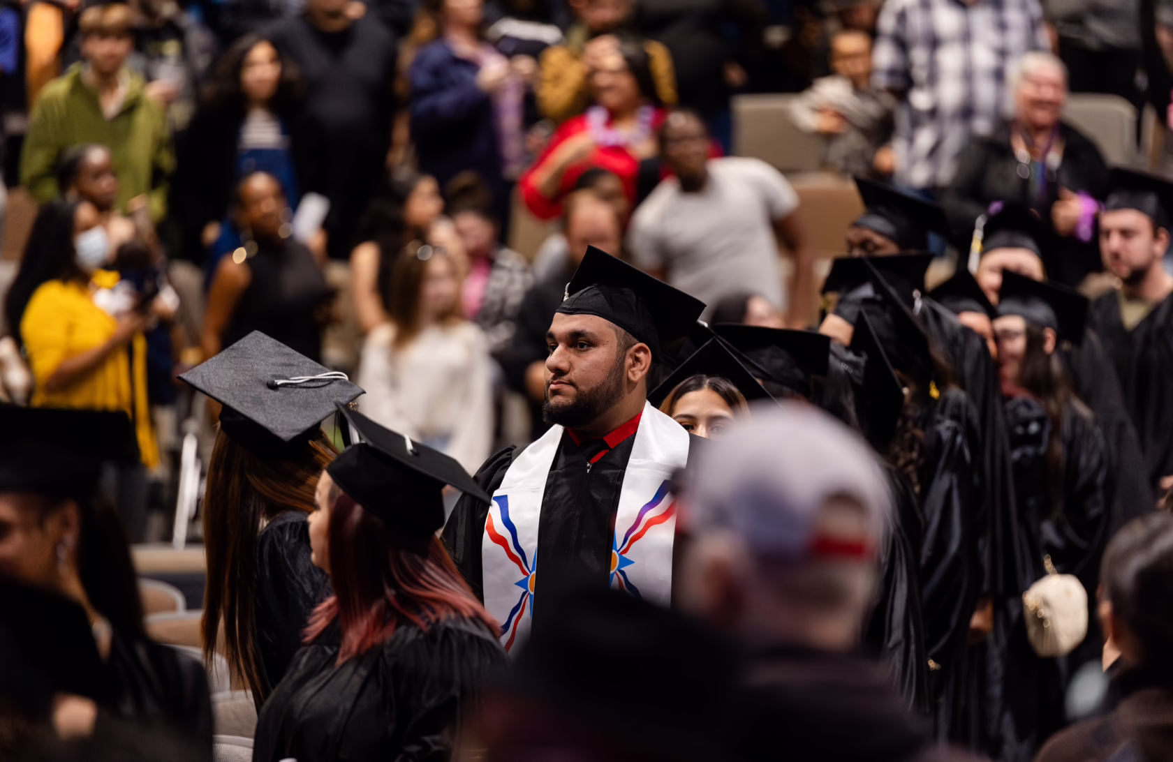 Graduates in black caps and gowns walk through an auditorium during a commencement ceremony, surrounded by audience members and fellow graduates.
