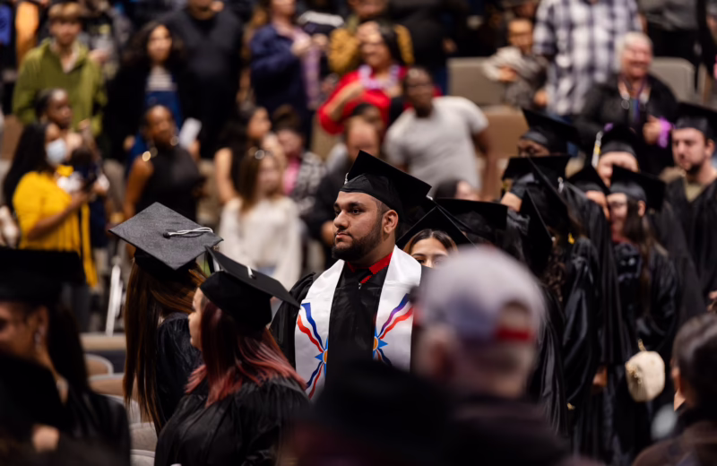 Graduates in black caps and gowns walk through an auditorium during a commencement ceremony, surrounded by audience members and fellow graduates.