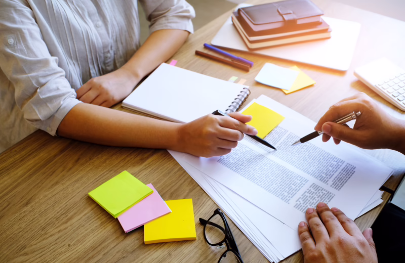 Two people reviewing printed documents at a desk, using pens and sticky notes to discuss and annotate materials.