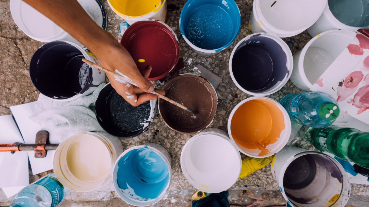 Overhead view of paint containers in multiple colors surrounding a hand holding a paintbrush during a painting project.