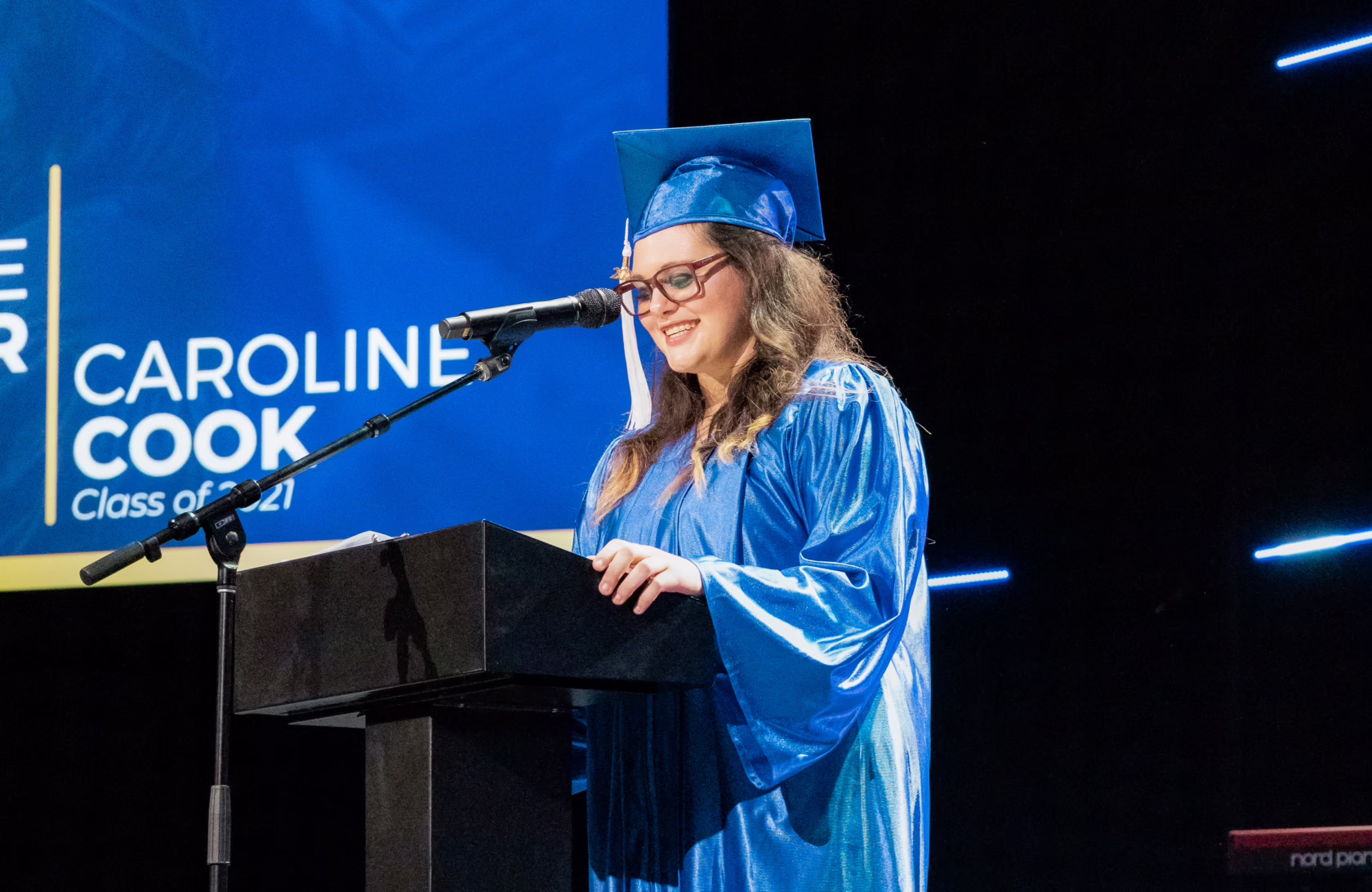 Graduate speaker in a blue cap and gown addresses the audience from a podium during a commencement ceremony, illuminated by blue stage lighting and speaking from prepared notes.